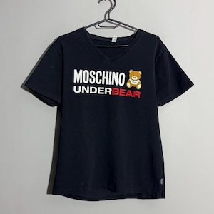 Puede incluir: Camiseta negra con cuello en V con el texto "MOSCHINO UNDERBEAR" en blanco y rojo. Un pequeño gráfico de oso de peluche de dibujos animados está en el lado derecho. La camiseta está colgada en una percha negra sobre un fondo gris.