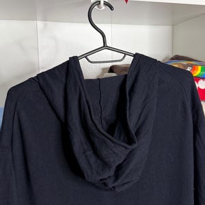 May include: A black hooded sweatshirt hanging on a black hanger. The hood is up, and the fabric appears to be a soft knit. The sweatshirt is a solid black color.