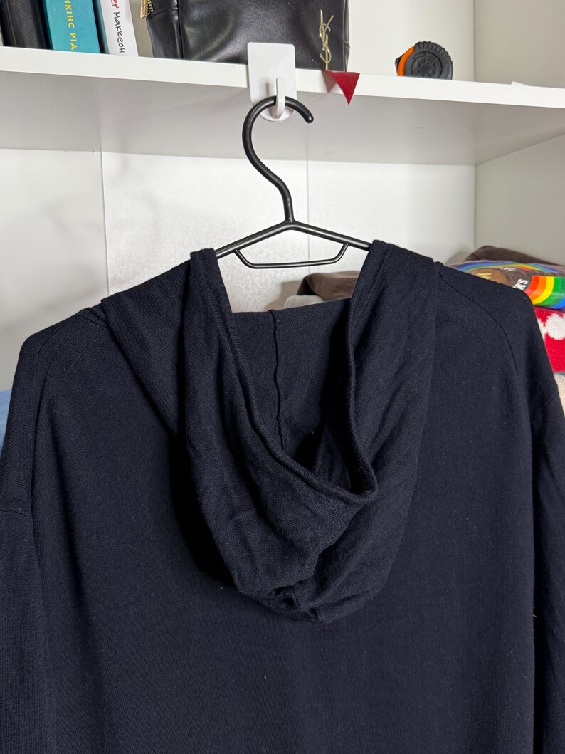May include: A black hooded sweatshirt hanging on a black hanger. The hood is up, and the fabric appears to be a soft knit. The sweatshirt is a solid black color.