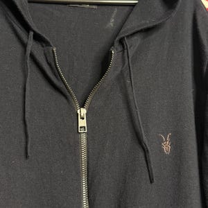 May include: A navy blue zip-up hoodie with a drawstring hood and a small embroidered logo on the chest. The hoodie has a full-length zipper and is hanging on a hanger.
