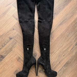 May include: A pair of black suede over-the-knee boots. The boots have a platform sole, stiletto heels, and zippers on the inner side. The boots are displayed on a wooden floor.