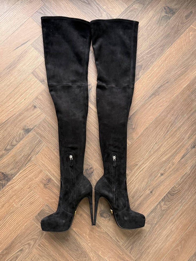 May include: A pair of black suede over-the-knee boots. The boots have a platform sole, stiletto heels, and zippers on the inner side. The boots are displayed on a wooden floor.