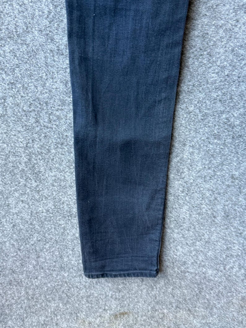 Op de afbeelding: Een enkele broekspijp gemaakt van donker antracietgrijze denimstof. Het materiaal heeft een zichtbare keperbinding en een recht model dat iets taps toeloopt naar de onderzoom. De stof vertoont een subtiele vervaging en een consistente donkere tint.
