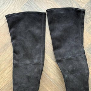 May include: A pair of black suede over-the-knee boots. The boots have a smooth, matte finish and a tall shaft that extends above the knee. The boots are displayed on a light brown wooden surface.