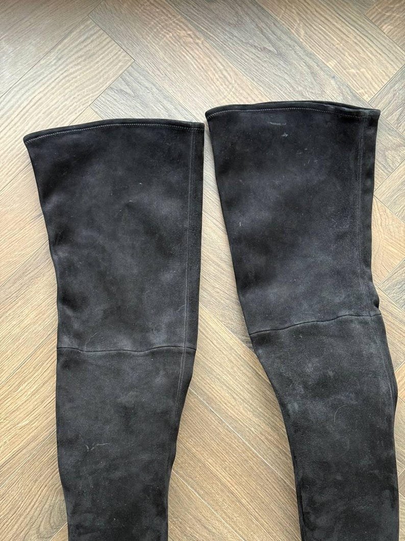 May include: A pair of black suede over-the-knee boots. The boots have a smooth, matte finish and a tall shaft that extends above the knee. The boots are displayed on a light brown wooden surface.
