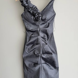 Puede incluir: Un vestido de c&oacute;ctel gris con cuello en V y un solo tirante adornado con volantes y una rosa de tela. El vestido tiene un corpi&ntilde;o fruncido y cuelga de una percha de madera.
