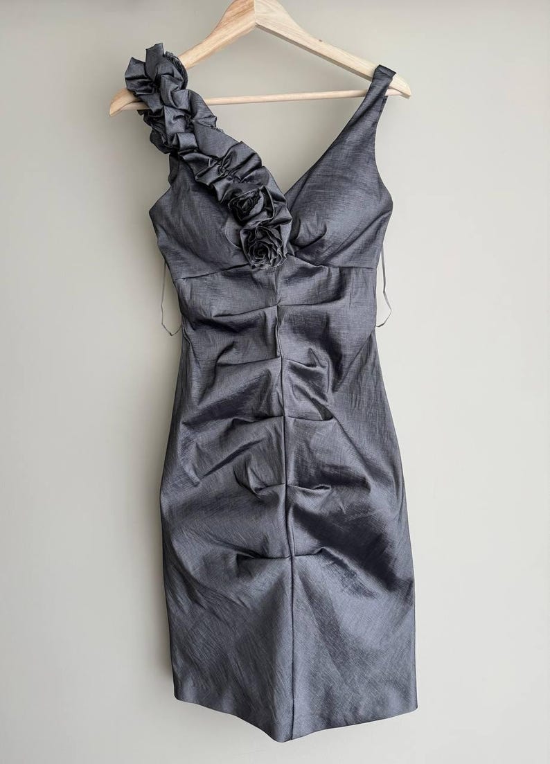 Puede incluir: Un vestido de c&oacute;ctel gris con cuello en V y un solo tirante adornado con volantes y una rosa de tela. El vestido tiene un corpi&ntilde;o fruncido y cuelga de una percha de madera.