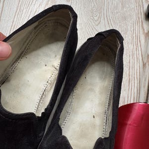 May include: Close-up of a pair of black suede slip-on shoes. The interior of the shoes is visible, showing the stitching and the brand name "Prada" on the side. The shoes appear worn.