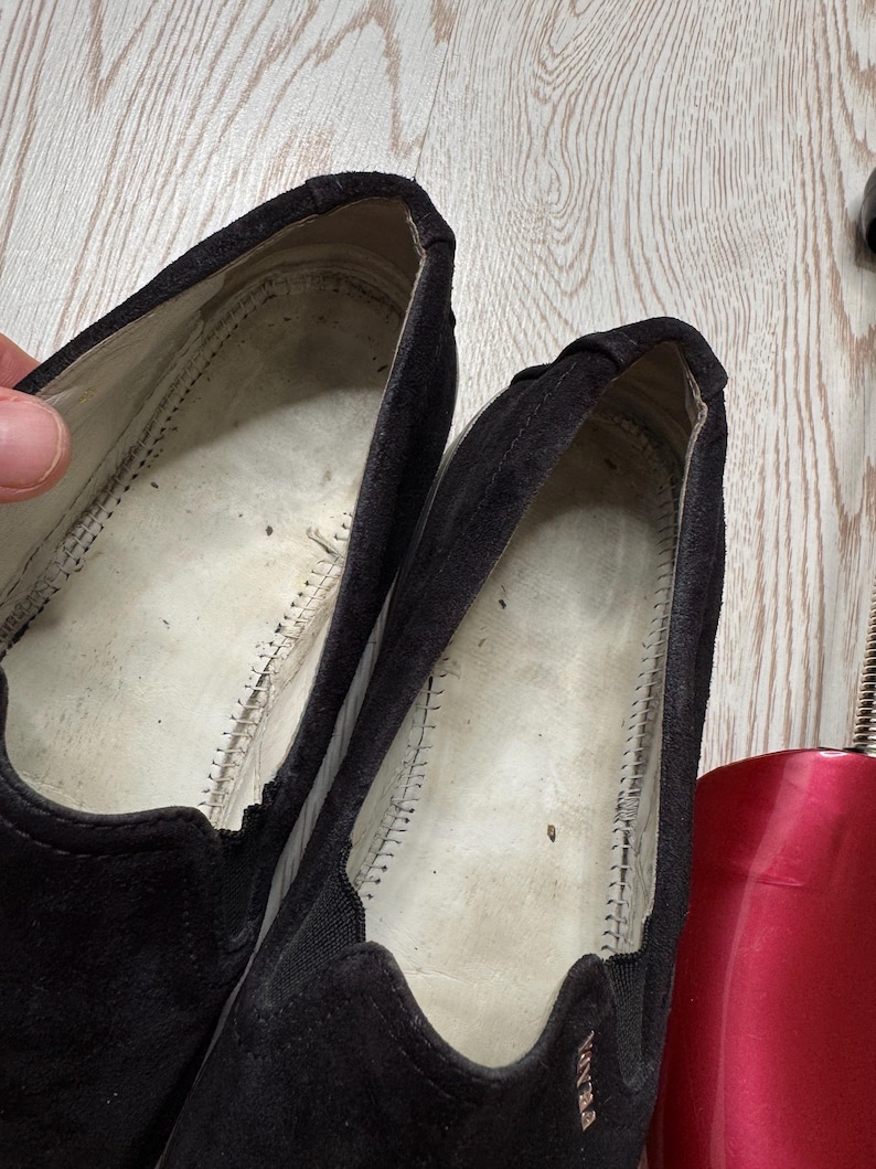 May include: Close-up of a pair of black suede slip-on shoes. The interior of the shoes is visible, showing the stitching and the brand name "Prada" on the side. The shoes appear worn.