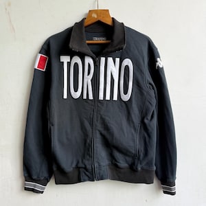 Può includere: Una giacca sportiva nera con zip e la scritta bianca TORINO sul petto. La giacca presenta un colletto alto, il logo del marchio Kappa all'interno del collo e sulla manica, e una piccola toppa quadrata rossa e bianca sulla manica destra.