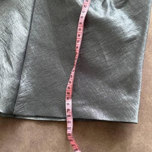 Puede incluir: Primer plano de una secci&oacute;n de tela gris con una cinta m&eacute;trica rosa. La cinta muestra medidas de 193 a 279 cm. La tela tiene un brillo sutil y parece ser un material liso.