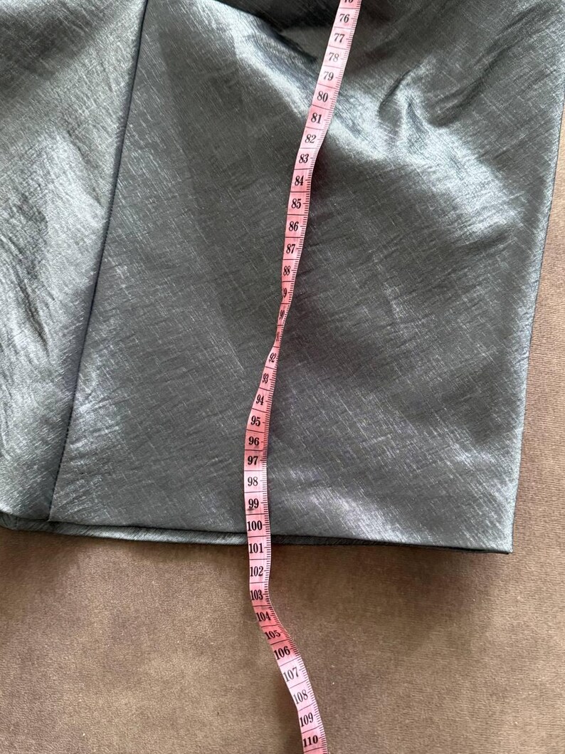 Puede incluir: Primer plano de una secci&oacute;n de tela gris con una cinta m&eacute;trica rosa. La cinta muestra medidas de 193 a 279 cm. La tela tiene un brillo sutil y parece ser un material liso.