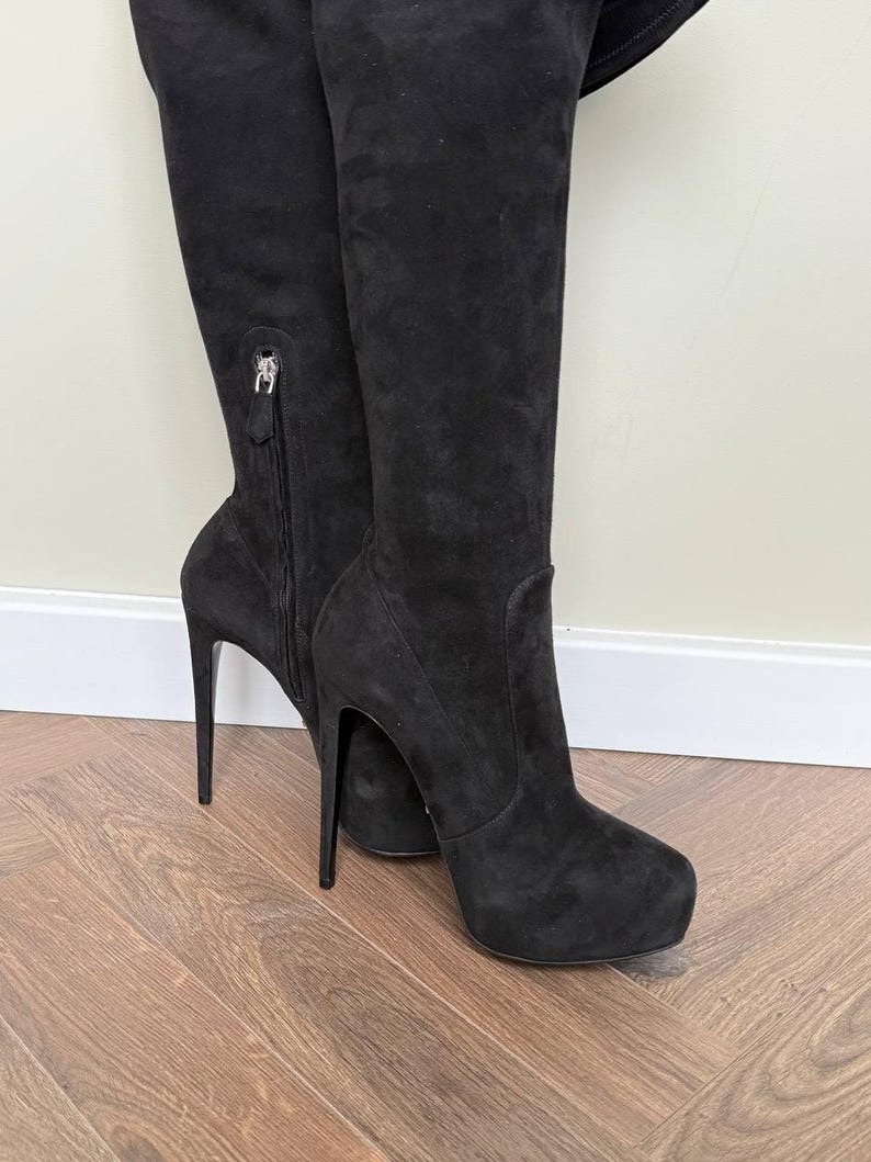 May include: Black suede over-the-knee boots with stiletto heels. The boots have a zipper detail on the side and a platform sole. The heels are approximately 5 inches (12.7 cm) high.