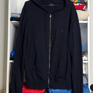 May include: A black zip-up hoodie with a hood and two front pockets hangs on a black hanger. The hoodie has a small logo on the chest. The background shows a white shelf with folded clothes.