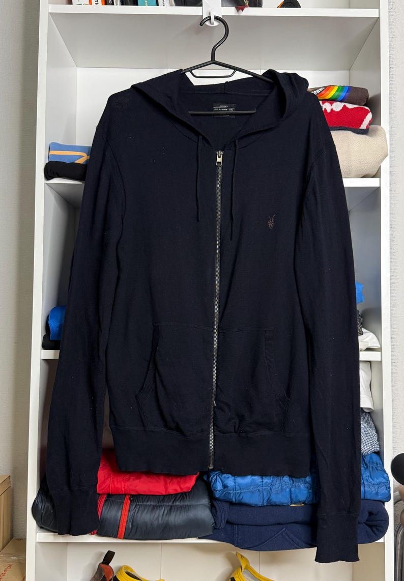May include: A black zip-up hoodie with a hood and two front pockets hangs on a black hanger. The hoodie has a small logo on the chest. The background shows a white shelf with folded clothes.