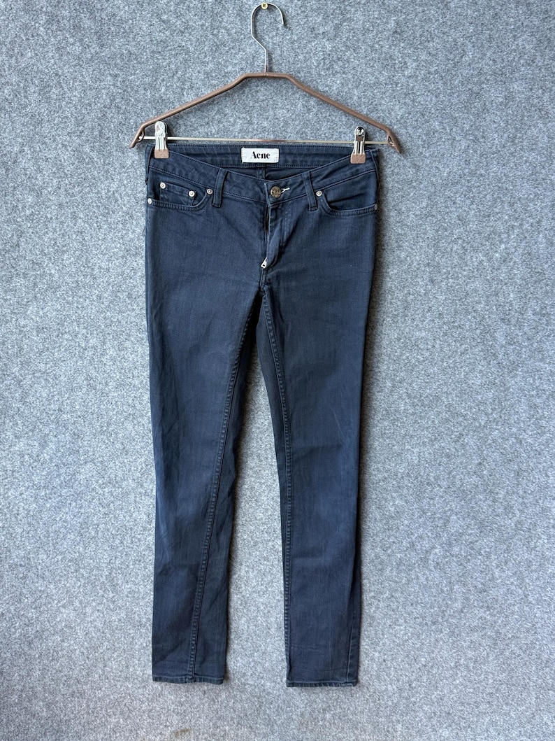 Op de afbeelding: Donkergrijze denim jeans met een klassiek five-pocket ontwerp, een knoop- en ritssluiting en een wit binnenlabel met de merknaam Acne in het zwart gedrukt, opgehangen aan een metalen kledinghanger met twee clips.