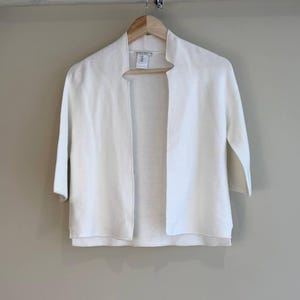 May include: A solid white open-front cardigan featuring a fine ribbed knit texture, three-quarter length sleeves, and a straight hemline. The garment is constructed with a clean, collarless neckline and a simple, unfastened front design.
