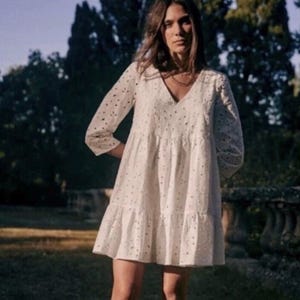 May include: A white, eyelet dress with a V-neck and three-quarter sleeves. The dress has a tiered skirt and is knee-length. The model is standing outside in a garden setting. The dress is a light, airy style.