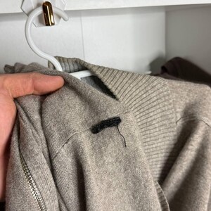 Pu&ograve; includere: Un maglione marrone chiaro con una cerniera sulla manica &egrave; appeso a una gruccia bianca. Il maglione ha un colletto a coste e una piccola etichetta nera sulla spalla. Lo sfondo &egrave; una mensola bianca.