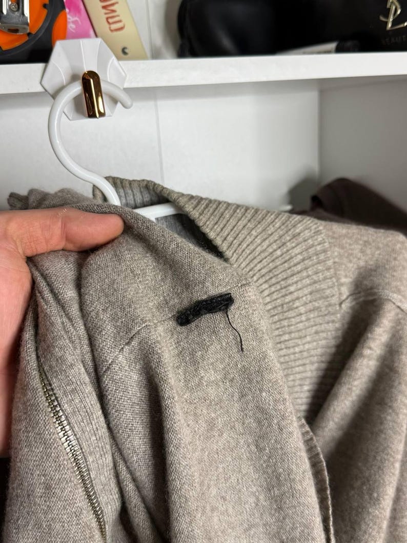 Pu&ograve; includere: Un maglione marrone chiaro con una cerniera sulla manica &egrave; appeso a una gruccia bianca. Il maglione ha un colletto a coste e una piccola etichetta nera sulla spalla. Lo sfondo &egrave; una mensola bianca.