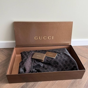 Op de afbeelding: Een bruine doos met het gouden woord "GUCCI" op het deksel. In de doos zit een donkere sjaal met een herhalend patroon en twee kleine kaartjes. De sjaal is opgevouwen en neemt het grootste deel van de doos in beslag.