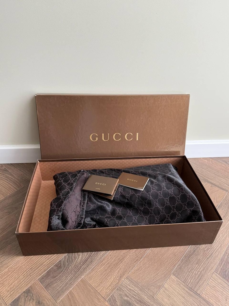 Op de afbeelding: Een bruine doos met het gouden woord "GUCCI" op het deksel. In de doos zit een donkere sjaal met een herhalend patroon en twee kleine kaartjes. De sjaal is opgevouwen en neemt het grootste deel van de doos in beslag.