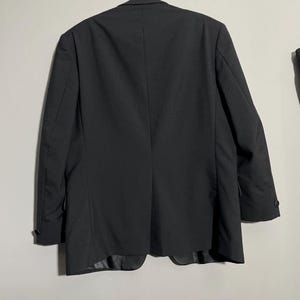 May include: A black blazer hanging on a metal hanger. The blazer has a classic design with a two-button closure and notched lapels. The back of the blazer is visible, showing the tailored fit and the vent at the bottom. The fabric appears to be a smooth, woven material.