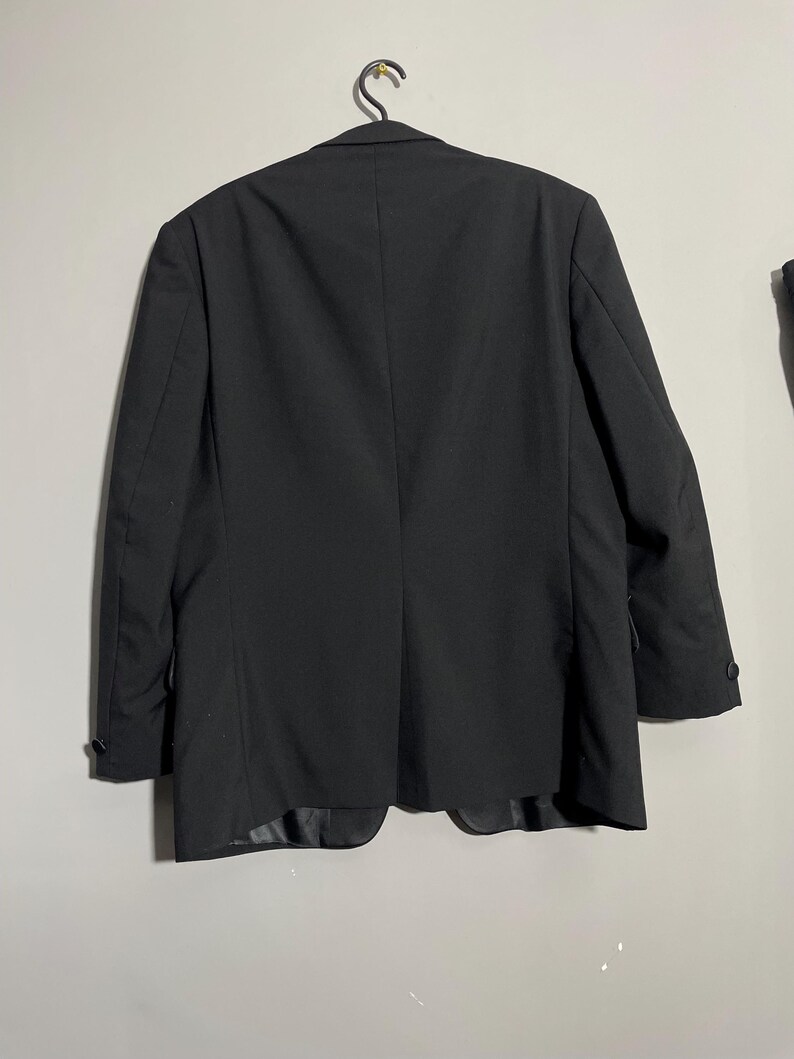 May include: A black blazer hanging on a metal hanger. The blazer has a classic design with a two-button closure and notched lapels. The back of the blazer is visible, showing the tailored fit and the vent at the bottom. The fabric appears to be a smooth, woven material.