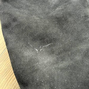 May include: Close-up of a black suede fabric. The material has a soft, textured appearance with subtle variations in color and a few small imperfections. The fabric is likely used for clothing or accessories.