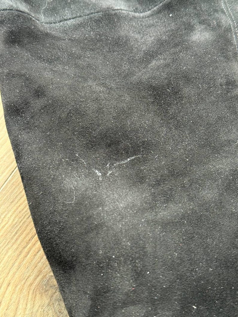May include: Close-up of a black suede fabric. The material has a soft, textured appearance with subtle variations in color and a few small imperfections. The fabric is likely used for clothing or accessories.