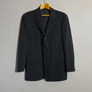 May include: A black blazer with a three-button fastening, suspended from a wooden hanger. The blazer features a notched lapel, two front pockets, and buttons on the sleeves. The garment is made of a smooth, dark fabric.