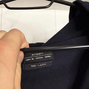 May include: A navy blue garment label with the brand name "ALLSAINTS" and the text "CONT. No - ORIGIN - CHINA". Below is the text "SIZE - LARGE". The label is attached to a navy blue garment.