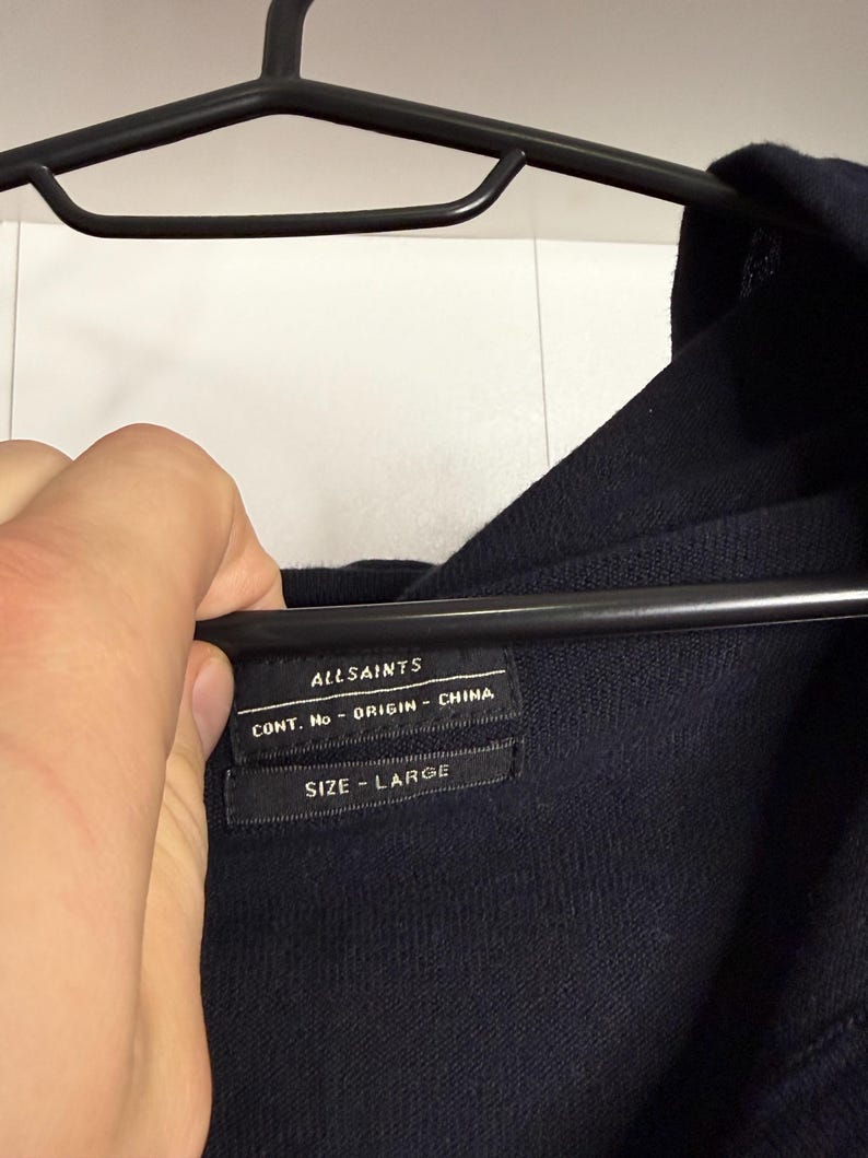May include: A navy blue garment label with the brand name "ALLSAINTS" and the text "CONT. No - ORIGIN - CHINA". Below is the text "SIZE - LARGE". The label is attached to a navy blue garment.