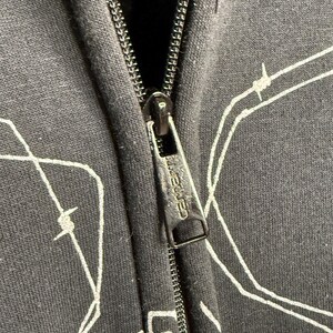 Pode incluir: Close-up de um moletom preto com z&iacute;per e um design de arame farpado branco. O puxador do z&iacute;per apresenta a marca "Carhartt". O moletom &eacute; feito de um tecido escuro.