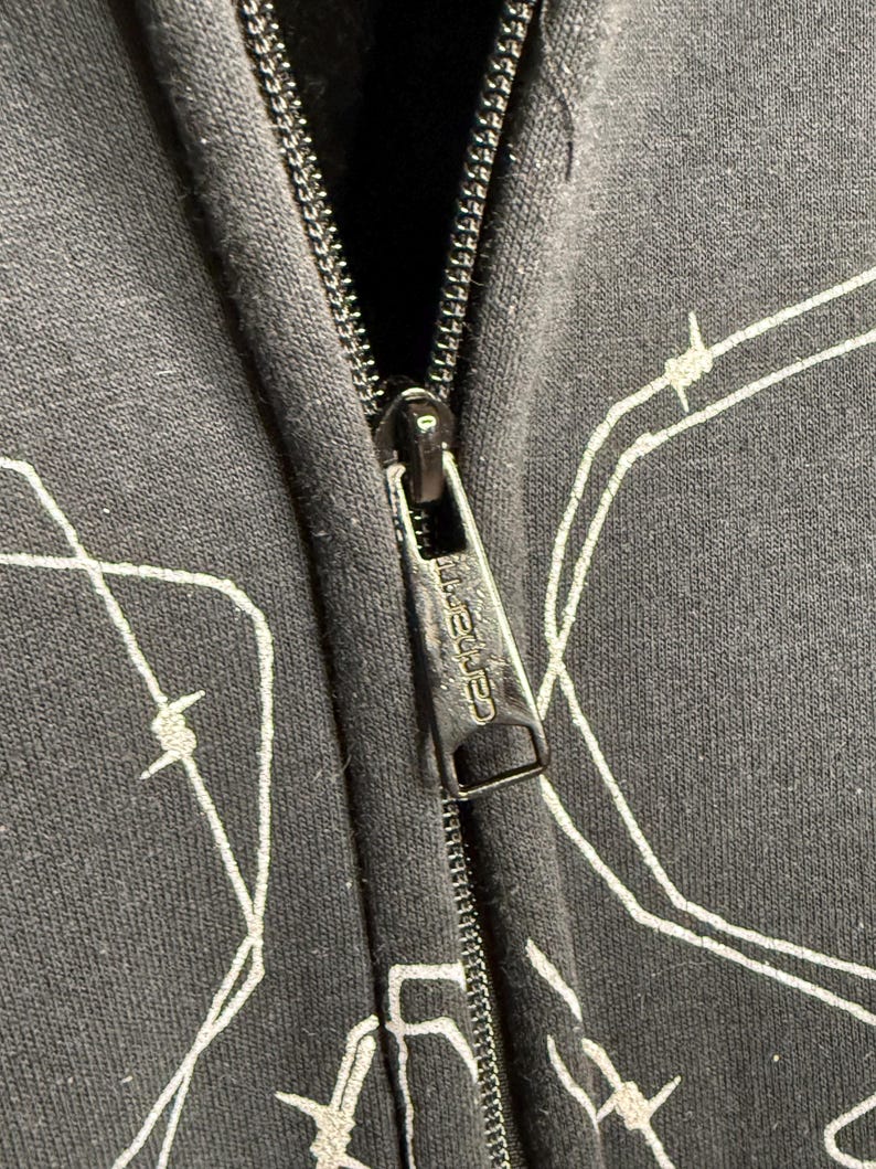 Pode incluir: Close-up de um moletom preto com z&iacute;per e um design de arame farpado branco. O puxador do z&iacute;per apresenta a marca "Carhartt". O moletom &eacute; feito de um tecido escuro.