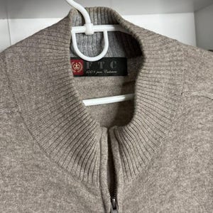 Pu&ograve; includere: Un maglione con zip marrone chiaro realizzato in 100% puro cashmere. Il maglione ha colletto e polsini a coste e una cerniera argentata. L'etichetta recita "FTC 100% pure Cashmere". Il maglione &egrave; appeso a una gruccia bianca.
