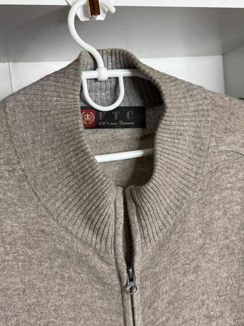 Pu&ograve; includere: Un maglione con zip marrone chiaro realizzato in 100% puro cashmere. Il maglione ha colletto e polsini a coste e una cerniera argentata. L'etichetta recita "FTC 100% pure Cashmere". Il maglione &egrave; appeso a una gruccia bianca.