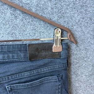 Op de afbeelding: Donkergrijze spijkerbroek van denim met een rechthoekige bruine leren patch op de achterkant van de tailleband, waarop het woord Acne in het midden is gedrukt. De broek wordt vastgehouden door een zilverkleurige metalen clip aan een bruine hanger.