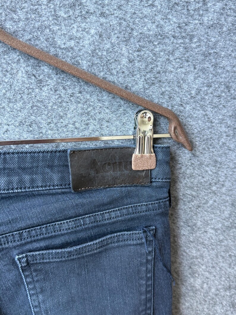 Op de afbeelding: Donkergrijze spijkerbroek van denim met een rechthoekige bruine leren patch op de achterkant van de tailleband, waarop het woord Acne in het midden is gedrukt. De broek wordt vastgehouden door een zilverkleurige metalen clip aan een bruine hanger.