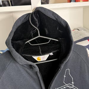Pode incluir: Moletom preto com z&iacute;per e capuz com um design gr&aacute;fico branco na frente. O moletom tem forro interno preto e uma etiqueta com o logotipo da Carhartt. O z&iacute;per &eacute; vis&iacute;vel no centro.