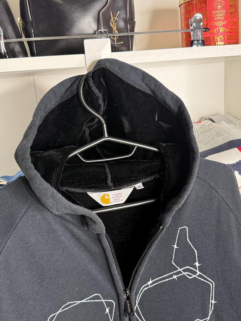 Pode incluir: Moletom preto com z&iacute;per e capuz com um design gr&aacute;fico branco na frente. O moletom tem forro interno preto e uma etiqueta com o logotipo da Carhartt. O z&iacute;per &eacute; vis&iacute;vel no centro.