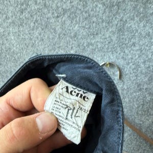 Op de afbeelding: Een donkergrijs denim kledingstuk met een wit stoffen label aan de binnenzijde waarop de merknaam Acne in het zwart is gedrukt. Het label toont maat 28/32 en bevat technische informatie, de materiaalsamenstelling en het land van herkomst.