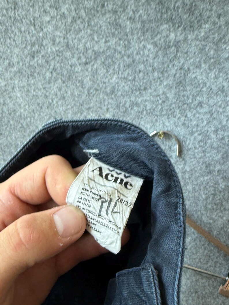 Op de afbeelding: Een donkergrijs denim kledingstuk met een wit stoffen label aan de binnenzijde waarop de merknaam Acne in het zwart is gedrukt. Het label toont maat 28/32 en bevat technische informatie, de materiaalsamenstelling en het land van herkomst.