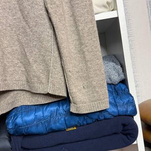 Pu&ograve; includere: Un maglione beige con bordi a coste &egrave; parzialmente visibile, insieme a una giacca trapuntata blu e una felpa blu navy. Gli articoli sono impilati all'interno di una mensola bianca.