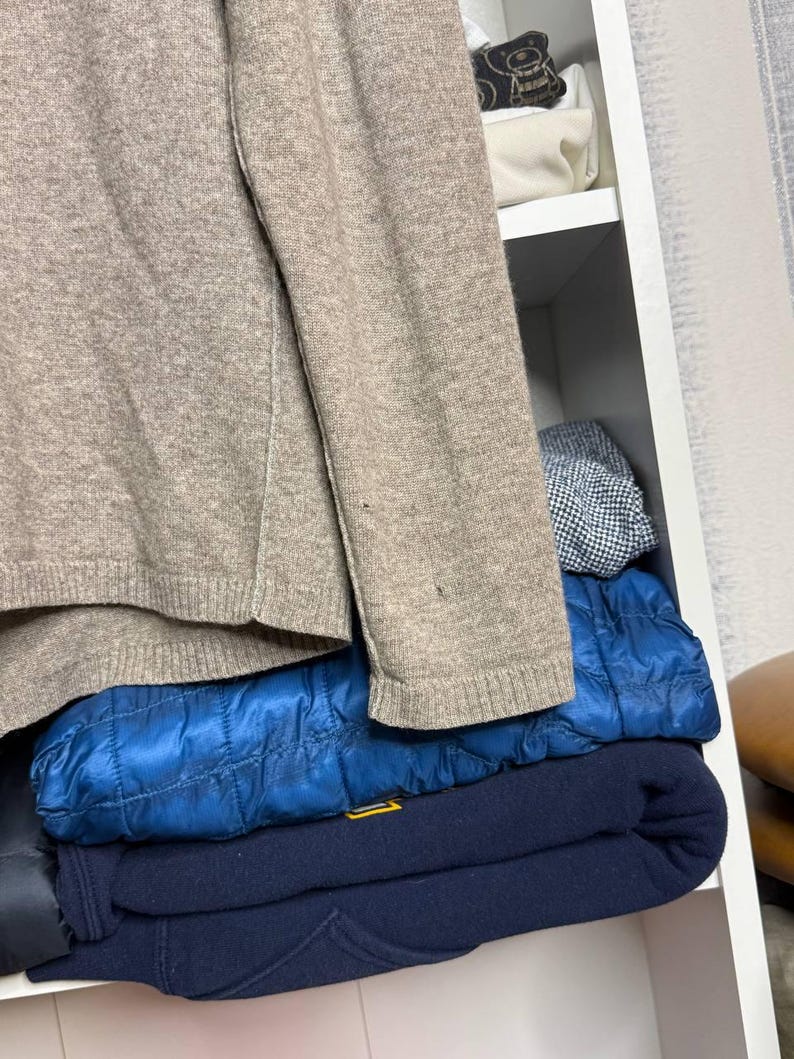 Pu&ograve; includere: Un maglione beige con bordi a coste &egrave; parzialmente visibile, insieme a una giacca trapuntata blu e una felpa blu navy. Gli articoli sono impilati all'interno di una mensola bianca.