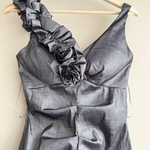 Puede incluir: Un vestido gris de un solo hombro con escote en V y un adorno floral decorativo. El vestido est&aacute; hecho de una tela brillante y cuelga de una percha de madera. La correa del hombro presenta tela con volantes.