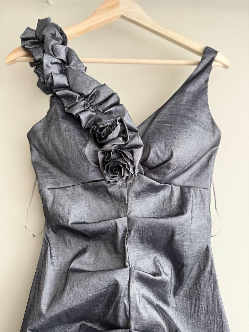 Puede incluir: Un vestido gris de un solo hombro con escote en V y un adorno floral decorativo. El vestido est&aacute; hecho de una tela brillante y cuelga de una percha de madera. La correa del hombro presenta tela con volantes.