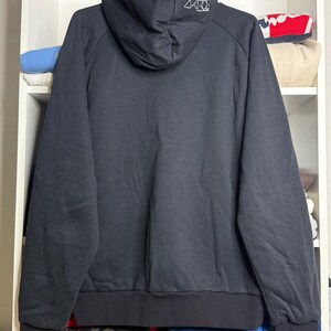 Pode incluir: Moletom preto com capuz com cord&atilde;o e bainha e punhos canelados. A parte de tr&aacute;s do capuz apresenta um design gr&aacute;fico branco. O moletom est&aacute; pendurado em um cabide preto.