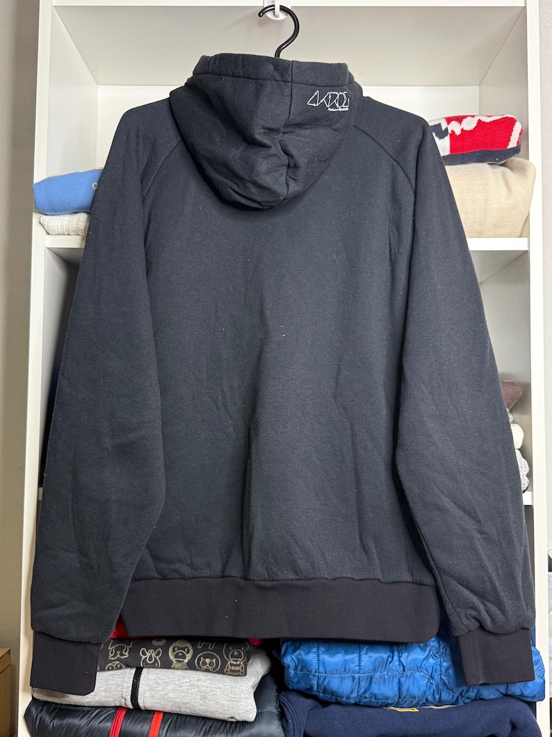 Pode incluir: Moletom preto com capuz com cord&atilde;o e bainha e punhos canelados. A parte de tr&aacute;s do capuz apresenta um design gr&aacute;fico branco. O moletom est&aacute; pendurado em um cabide preto.