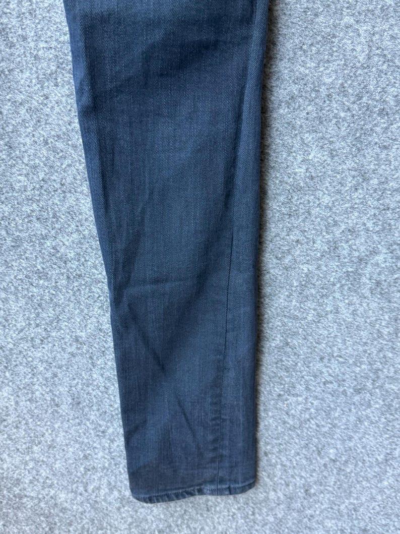 Op de afbeelding: Een enkele broekspijp gemaakt van donker antracietgrijze denimstof. Het materiaal heeft een zichtbare keperbinding en een recht model. Het kledingstuk is van bovenaf gefotografeerd, waardoor de lengte en de textuur van de stof zichtbaar zijn.
